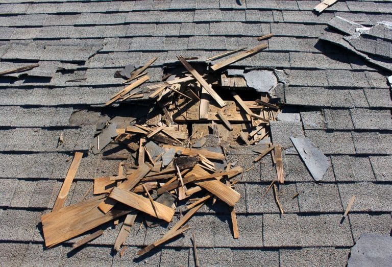 Close-up of a severely damaged shingle roof with a large hole exposing the underlying wood structure and scattered broken roofing materials.